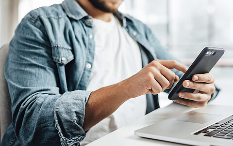 Mann sitzt an einem Tisch mit Notebook und bedient ein Smartphone